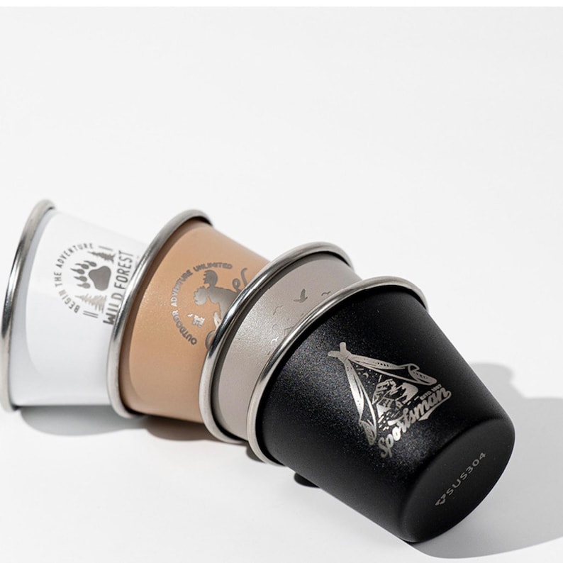 May include: Four stainless steel camping cups in white, tan, grey, and black, stacked together. Each cup has a silver rim and a different outdoor-themed graphic. The white cup says "Wild Forest" and the black cup says "Sportsman".