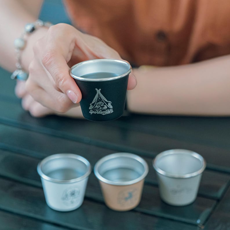 May include: A black metal cup with a silver rim, held by a person. The cup features a camping tent graphic and the word "Sportsman". Three additional metal cups in white, tan, and silver are visible below.