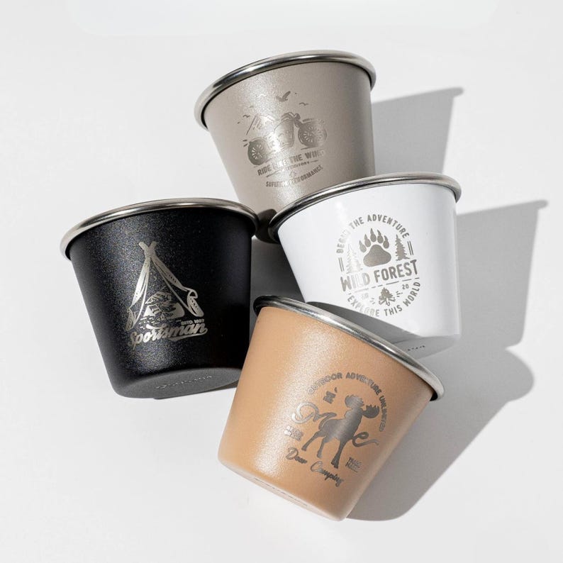 May include: Four metal camping cups in black, tan, white, and grey. Each cup has a silver rim and a unique graphic design, including a motorcycle, a tent, a bear paw, and a moose. The cups are stacked on a white surface.