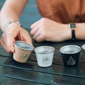 May include: A collection of small, metal camping cups in beige, white, and black. Each cup has a different outdoor-themed graphic and text. The cups are designed for outdoor adventures and camping.