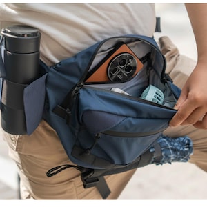 May include: A navy blue bum bag with several compartments, including a water bottle holder and a zipped main pocket. The main pocket contains a phone with a brown case, a small fan, and other items. A black water bottle is in the side holder.