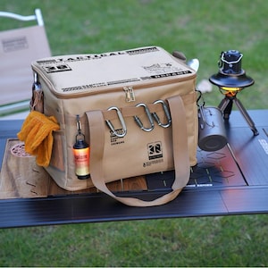 May include: A tan camping cooler bag with the text "TACTICAL CAMPING" and a zip closure. The bag has a shoulder strap, multiple carabiners, and a side pocket with orange gloves. A small lantern hangs from the bag.