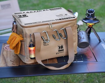 Può includere: Una borsa frigo da campeggio beige con la scritta "TACTICAL CAMPING" e chiusura a zip. La borsa ha una tracolla, diversi moschettoni e una tasca laterale con guanti arancioni. Una piccola lanterna è appesa alla borsa.