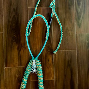 Puede incluir: Un cabestro de caballo de cuerda turquesa y beige con un diseño trenzado. El cabestro presenta un lazo para la cabeza del caballo y una sección para la nariz, con una borla decorativa en la parte inferior. La cuerda es una combinación de turquesa y beige.