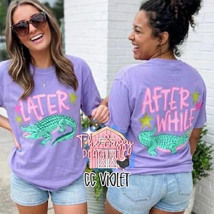 May include: Two women wearing lavender t-shirts with a green alligator graphic. The front of the shirt says "LATER" and the back says "AFTER WHILE" in pink lettering. The shirts are paired with denim shorts.