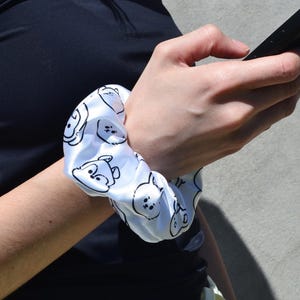 May include: A white fabric scrunchie featuring a repeating black line art pattern of cartoon animal faces. The accessory has a smooth, slightly reflective surface and a gathered, elasticized construction designed to hold hair securely in place.