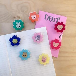 May include: A collection of colorful flower-shaped magnets with animal faces, including a bunny, bear, and cat. The magnets are on a notebook and a pink paper with the word "Doit".