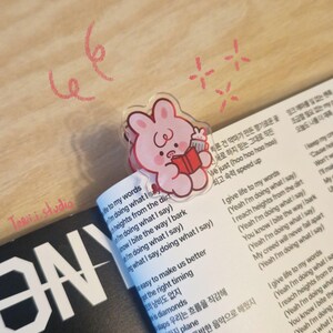 May include: A clear acrylic bookmark featuring a pink cartoon bunny holding a book and a drink, placed on a book with Korean text. The bookmark is on a page with English lyrics. The words "Torii studio" are visible.
