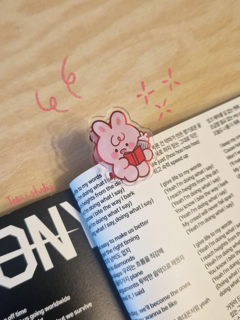May include: A clear acrylic bookmark featuring a pink cartoon bunny holding a book and a drink, placed on a book with Korean text. The bookmark is on a page with English lyrics. The words "Torii studio" are visible.
