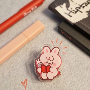 May include: A clear acrylic pin featuring a pink cartoon bunny reading a red book and holding a cupcake. A red pen and a peach-colored highlighter are in the background. The pin has a red outline and is on a gray surface.