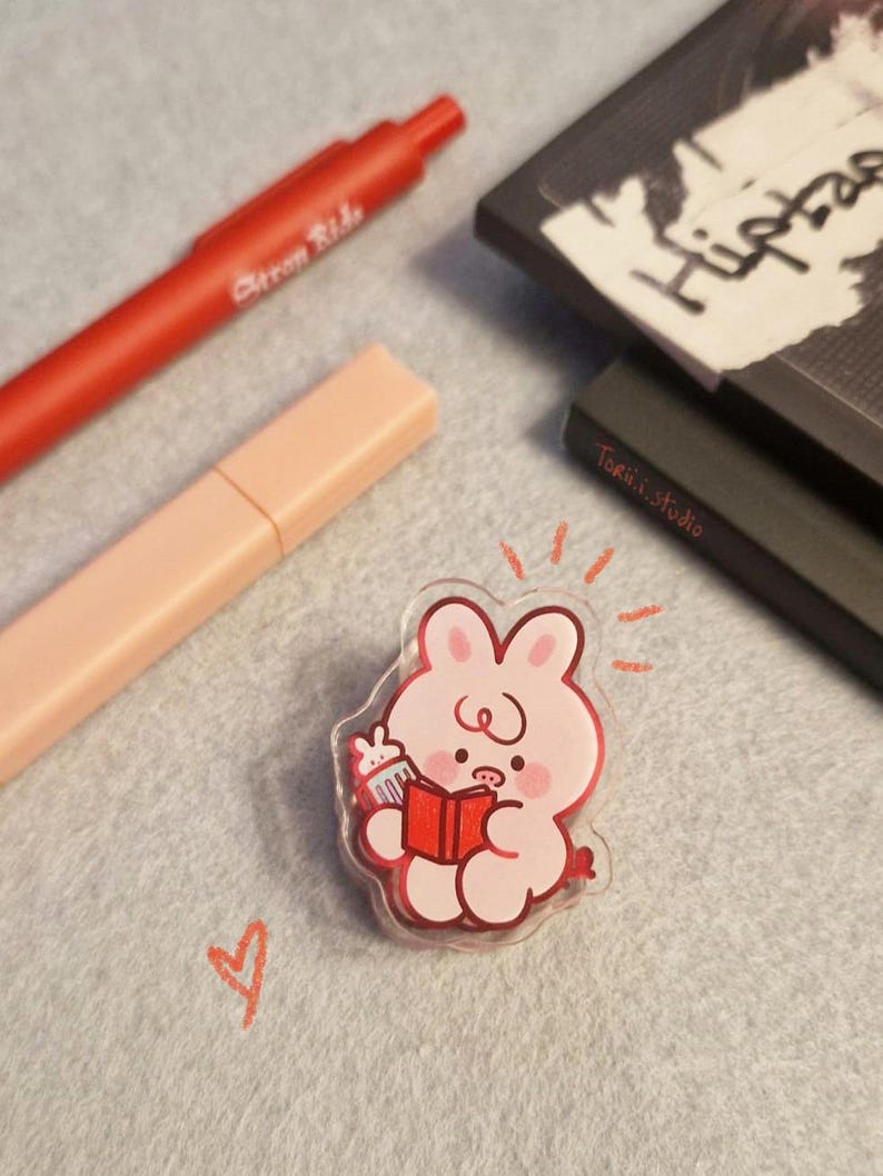 May include: A clear acrylic pin featuring a pink cartoon bunny reading a red book and holding a cupcake. A red pen and a peach-colored highlighter are in the background. The pin has a red outline and is on a gray surface.