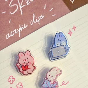 May include: Three acrylic clips featuring cartoon bunnies in various poses. One pink bunny reads a book, another blue bunny works on a laptop, and a third white bunny rides a scooter. The text "SKzoo acrylic clips" is written in white.