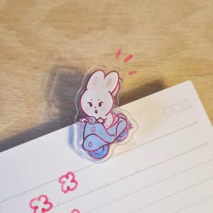 May include: A clear acrylic bunny riding a blue scooter. The bunny is pink and white, with a surprised expression. The scooter has blue wheels and accents. The image is on a white lined paper with pink flower doodles and the text "Torii.i.studio".