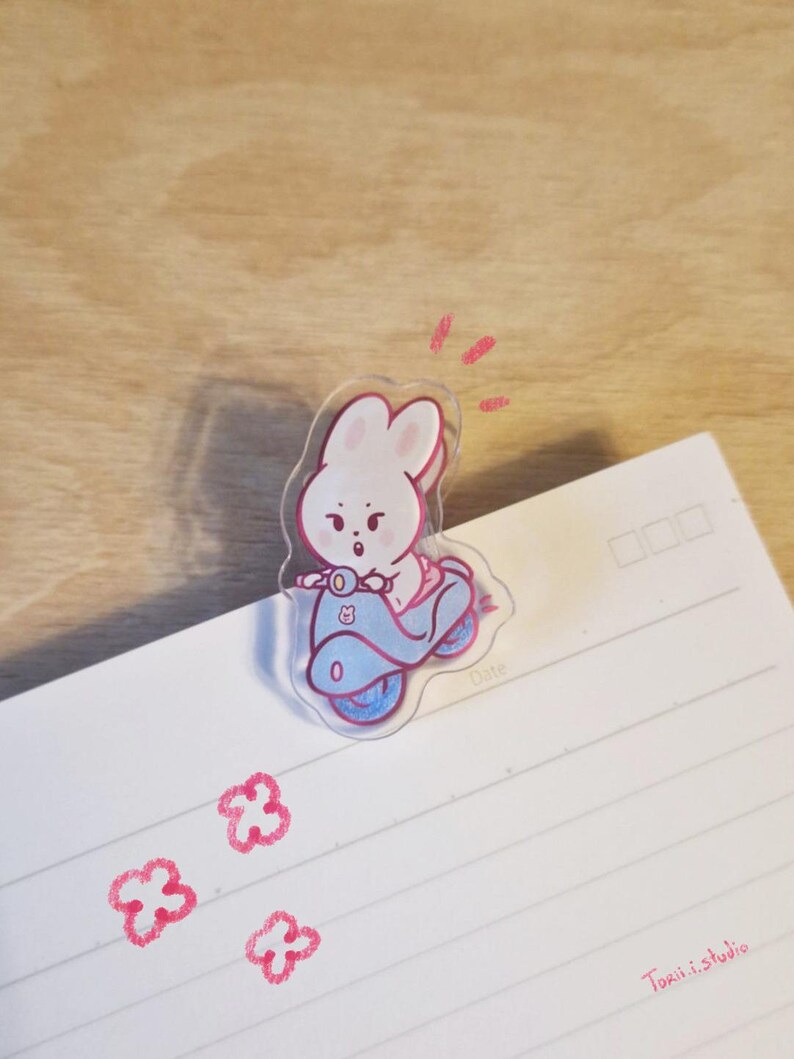 May include: A clear acrylic bunny riding a blue scooter. The bunny is pink and white, with a surprised expression. The scooter has blue wheels and accents. The image is on a white lined paper with pink flower doodles and the text "Torii.i.studio".