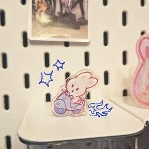 May include: A clear acrylic pin featuring a cartoon bunny riding a light blue scooter. The bunny is pink and white. A small photo of a group of people is in the background. The pin is on a white shelf.