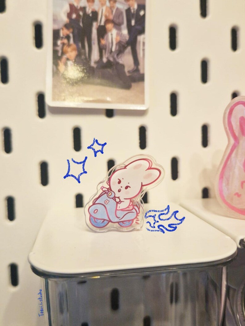 May include: A clear acrylic pin featuring a cartoon bunny riding a light blue scooter. The bunny is pink and white. A small photo of a group of people is in the background. The pin is on a white shelf.