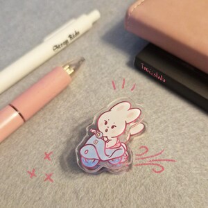 May include: A clear acrylic sticker featuring a cartoon bunny riding a light blue scooter with pink accents. The bunny is wearing a pink outfit. The sticker is on a gray surface with a pink pen and a white pen.