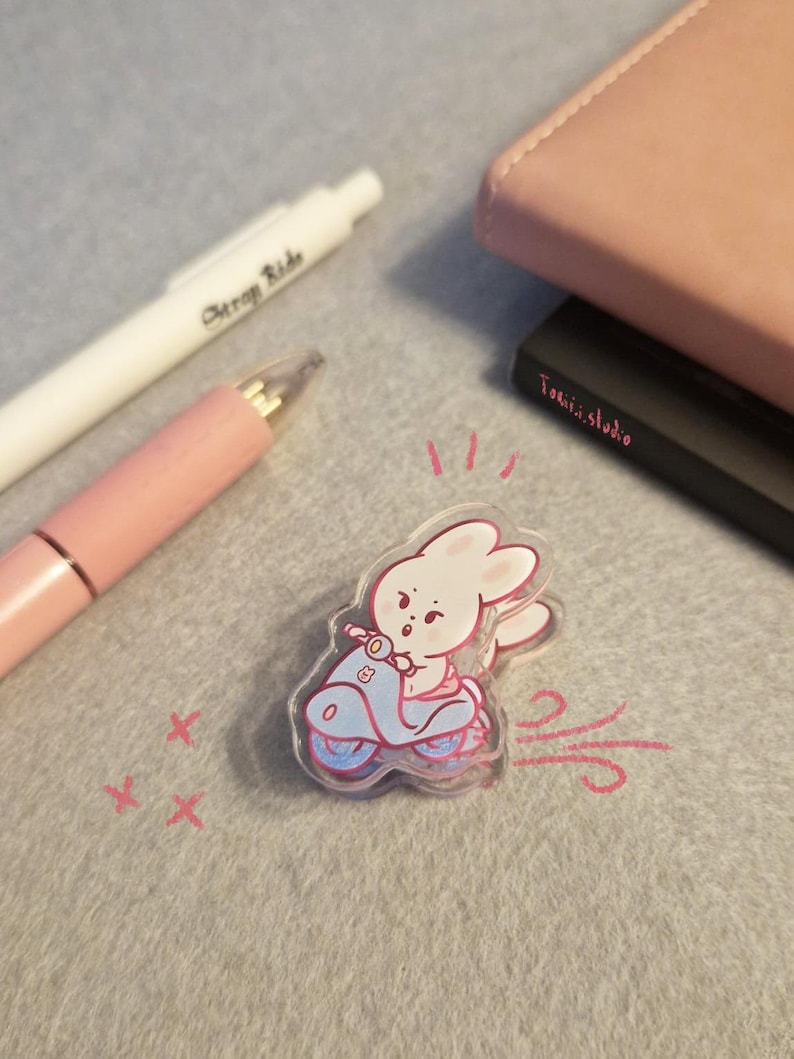 May include: A clear acrylic sticker featuring a cartoon bunny riding a light blue scooter with pink accents. The bunny is wearing a pink outfit. The sticker is on a gray surface with a pink pen and a white pen.