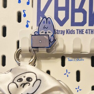 May include: A white pegboard with a clear acrylic charm of a blue cartoon character using a laptop. The charm is attached to a white hook. A white and black patterned scarf hangs below. Blue musical notes and crosses are painted on the board.