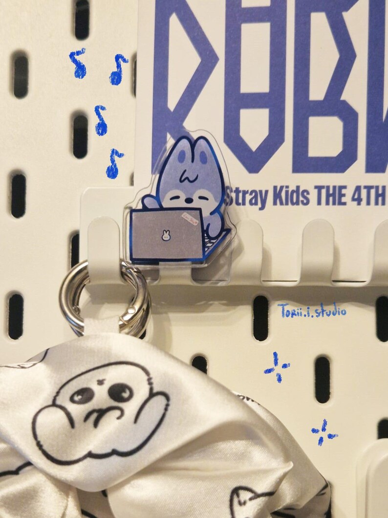 May include: A white pegboard with a clear acrylic charm of a blue cartoon character using a laptop. The charm is attached to a white hook. A white and black patterned scarf hangs below. Blue musical notes and crosses are painted on the board.