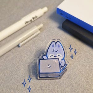 May include: A clear acrylic pin featuring a blue cartoon bunny working on a laptop. The pin has a blue outline and is decorated with blue star shapes. Two white pens and a notebook are in the background.