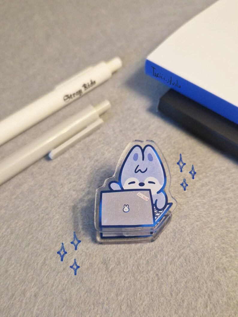 May include: A clear acrylic pin featuring a blue cartoon bunny working on a laptop. The pin has a blue outline and is decorated with blue star shapes. Two white pens and a notebook are in the background.