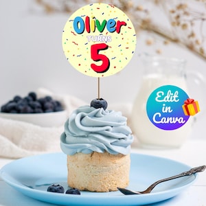 May include: A blue cupcake with white frosting and a single blueberry on top. A yellow and white circular cake topper with the text "Oliver turns 5" in colorful letters.
