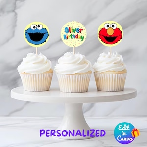 May include: Three white cupcakes with white frosting on a white cake stand. Each cupcake has a different Sesame Street character topper: Cookie Monster, Elmo, and a yellow circle with the text "Oliver Birthday".