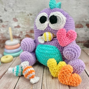 Puede incluir: Un juguete de peluche de ganchillo hecho a mano con forma de monstruo morado con grandes ojos con borde blanco y una panza turquesa. Tiene corazones de ganchillo en rosa, amarillo y naranja, y juguetes con forma de caramelo. Fondo de madera.