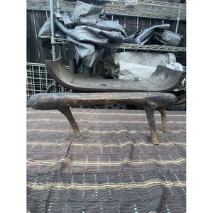 Puede incluir: Un banco rústico de madera con tres patas y una parte superior curvada, junto con un abrevadero a juego. El banco y el abrevadero están hechos de madera oscura y desgastada, mostrando la veta y la textura naturales. Los artículos están colocados sobre una tela estampada.
