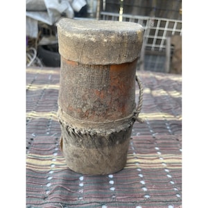 Op de afbeelding: Een rustieke, cilindrische houten container met een verweerde uitstraling. De container heeft een deksel en is gebonden met touw. Het hout vertoont zichtbare nerf- en kleurvariaties, wat duidt op leeftijd en gebruik. De container is waarschijnlijk voor opslag.