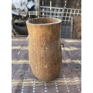 May include: A tall, cylindrical wooden vase with a rough, textured surface and a natural brown color. The vase has two small, rounded handles and a wide opening. It is placed on a patterned fabric.