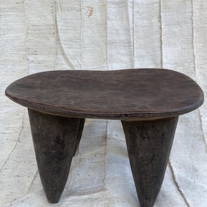 May include: A dark brown wooden stool featuring a smooth, irregularly shaped oval seat supported by three thick, tapered legs. The wood displays a natural, weathered texture with visible grain patterns throughout the entire structure of the piece.