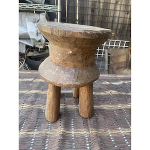 May include: A small, handcrafted wooden stool with a round top and three sturdy legs. The stool has a natural, aged appearance with visible wood grain and a warm, brown color. It is likely an antique or vintage piece.