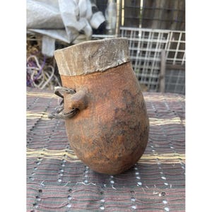 Puede incluir: Un recipiente artesanal en forma de calabaza, de color marrón, con un borde claro y un asa pequeña. El recipiente tiene una superficie texturizada y parece estar hecho de materiales naturales. La calabaza está sobre una superficie estampada.
