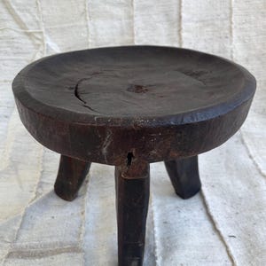 May include: A dark brown, circular wooden stool featuring a thick, flat top with a visible natural crack and three sturdy, carved legs. The surface of the wood shows a weathered, textured finish with deep grain patterns and signs of age.