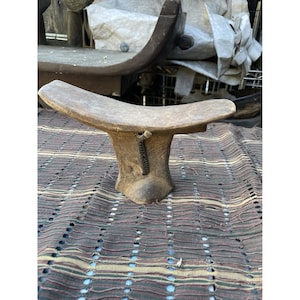 Puede incluir: Un reposacabezas de madera desgastada con una parte superior curvada y una base ensanchada. El reposacabezas es de color marrón claro y parece hecho a mano. Un detalle oscuro con cuentas cuelga del lateral. El reposacabezas está colocado sobre un textil estampado.