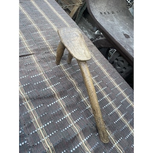 Puede incluir: Un taburete rústico de madera de tres patas con un mango largo. El taburete está hecho de madera de color claro y tiene un aspecto desgastado y envejecido. El taburete está sobre un fondo estampado.