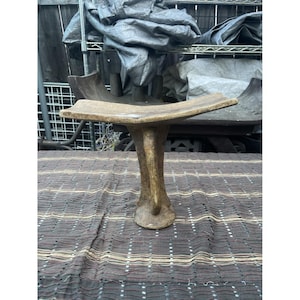 Puede incluir: Un taburete de madera rústico y artesanal con una sola pata robusta. El taburete tiene un aspecto natural y envejecido con un asiento plano y rectangular. La madera es de color marrón claro y parece estar hecha de una sola pieza.