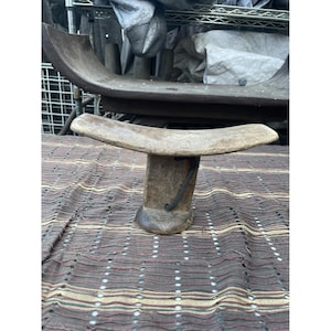 Puede incluir: Un reposacabezas de madera con una parte superior curvada y una base cilíndrica. El reposacabezas está hecho de madera de color claro y tiene un acento de metal oscuro. Se encuentra sobre un textil estampado.