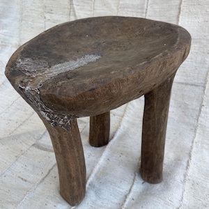 May include: A small, three-legged stool carved from dark brown wood. The seat features a prominent, irregular crack filled with a metallic, silver-toned material that extends from the edge toward the center of the textured, weathered surface.