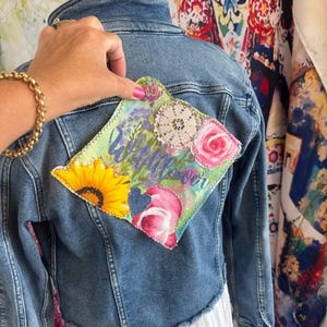 Può includere: Giacca di jeans blu con una toppa decorativa sul retro. La toppa presenta un girasole, rose e la parola "Wildflower" in corsivo. La giacca ha una vestibilità corta e un vestito bianco è visibile sotto.