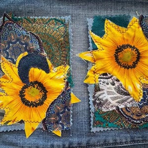 Op de afbeelding: Twee stoffen patches met zonnebloemontwerpen. De zonnebloemen zijn geel met donkere centra en gestikte randen. De patches bevinden zich op een denim achtergrond, met patronen van blauw, groen en goud.