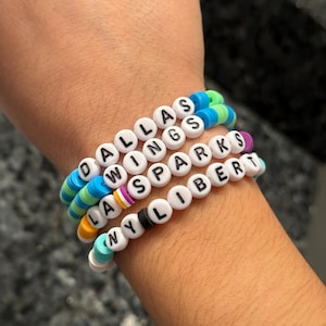 Puede incluir: Un conjunto de pulseras de cuentas de colores con las palabras "Dallas Wings", "LA Sparks", "NY Liberty" y "Liber" escritas en letras negras.