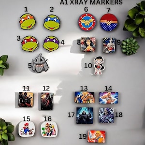 May include: A1 XRAY MARKERS, a set of 19 colourful, square and round markers featuring various cartoon characters, including Teenage Mutant Ninja Turtles, Wonder Woman, Deadpool, He-Man, and Super Mario.