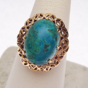 May include: Gold-toned ring featuring a large, oval-shaped gemstone with a vibrant teal and green color. The gemstone is set within a decorative, openwork gold filigree setting, creating an elegant and eye-catching design.