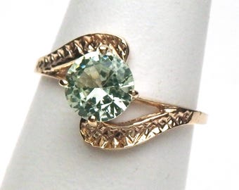 May include: A gold-toned ring featuring a large, round, light green gemstone. The ring's band has a textured, swirling design that curves around the gemstone. The gemstone is held in place by four prongs.