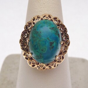 May include: A gold-toned ring featuring a large, oval-shaped gemstone with a vibrant teal and green color. The gemstone is set within a decorative, openwork gold frame. The ring is displayed on a white stand.