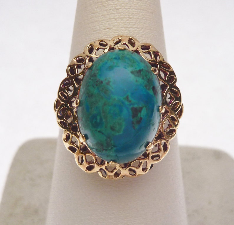 May include: A gold-toned ring featuring a large, oval-shaped gemstone with a vibrant teal and green color. The gemstone is set within a decorative, openwork gold frame. The ring is displayed on a white stand.