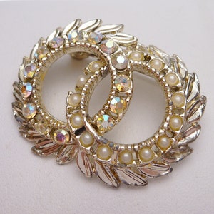 Puede incluir: Un broche de tono plateado con dos círculos entrelazados decorados con piedras de imitación de cristal de colores iridiscentes y perlas falsas.